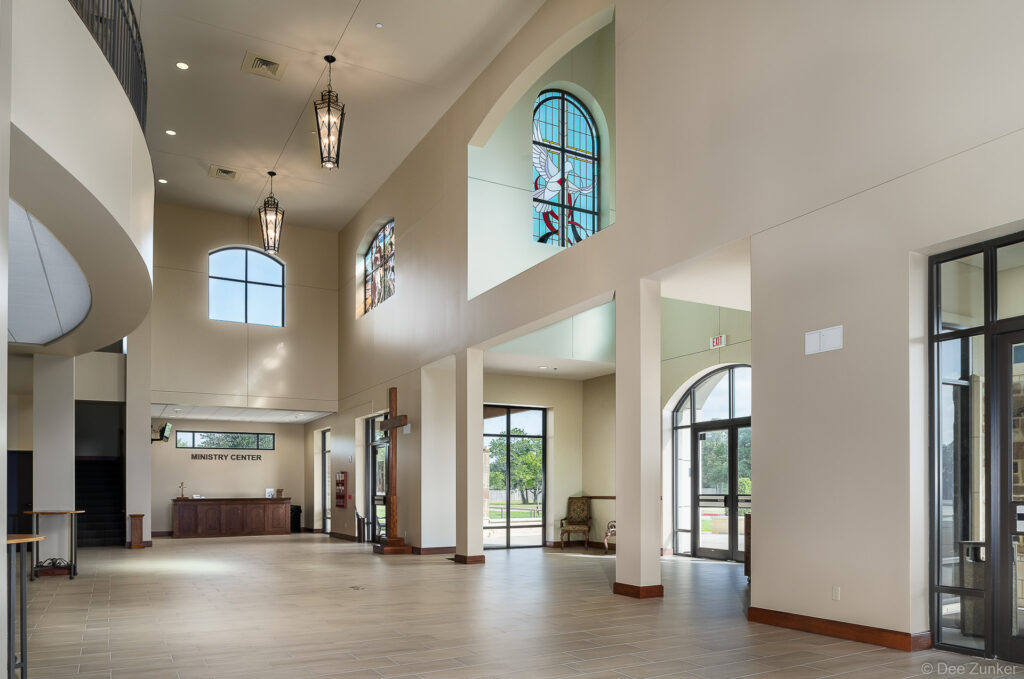 Soaring church entryway lobby with stained glass dove window, pendant chandeliers, arched windows, and ministry center reception desk