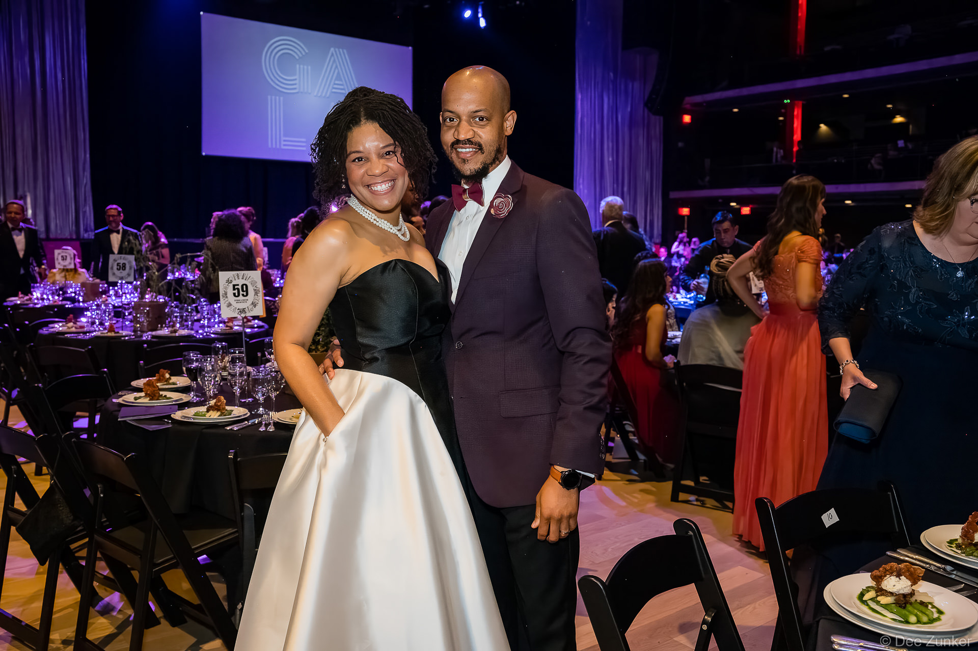 Smiling couple poses at Gala Houston corporate event, woman in black and white gown, man in burgundy suit with bow tie.