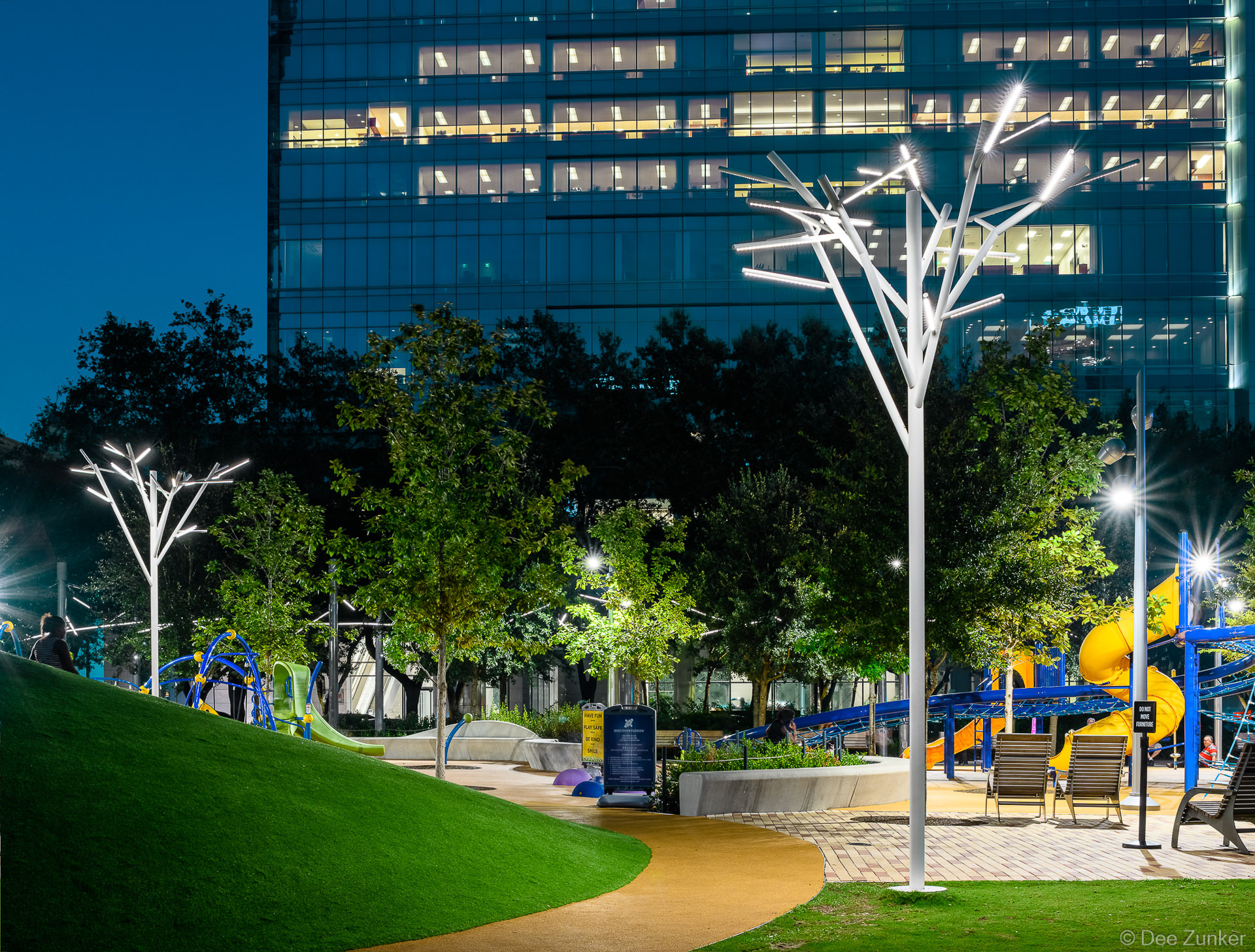 Tree-shaped LED light sculptures illuminating Discovery Green Park at night with colorful playground equipment and downtown Houston skyscraper backdrop