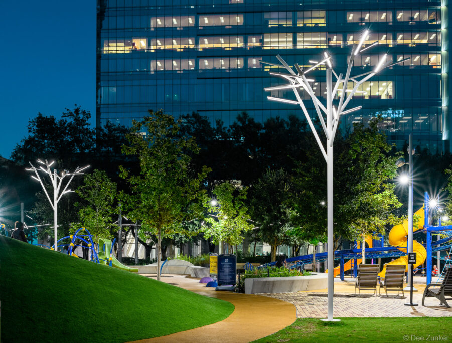 Tree-shaped LED light sculptures illuminate Discovery Green park at night with colorful playground equipment and Houston skyline behind