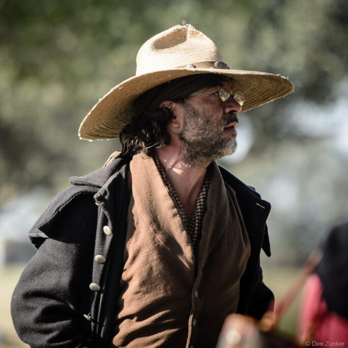 Bearded male reenactor in worn straw hat and period frontier clothing at George Ranch Historical Park, Houston area event photography.