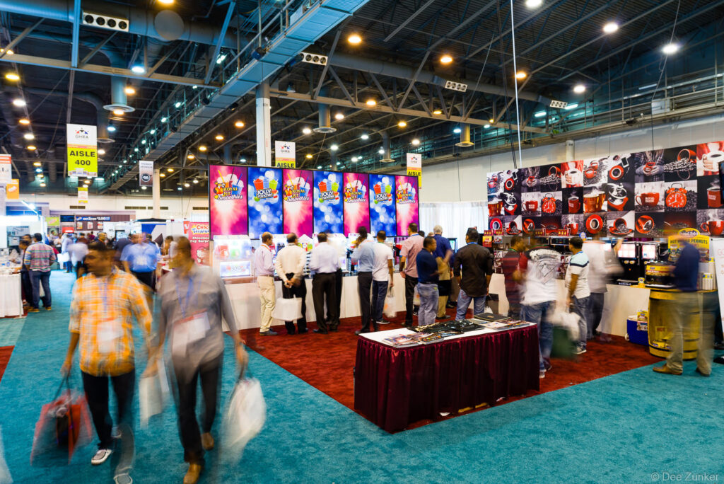 Busy trade show convention floor with vendor booths, colorful display screens, and attendees browsing exhibits at a Houston industry event.