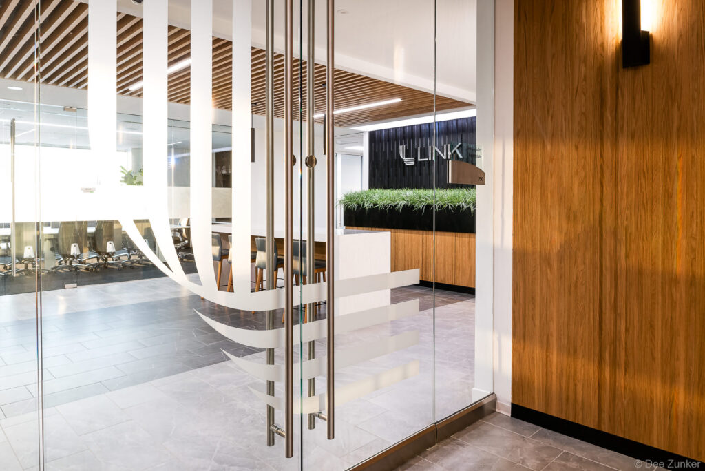 Glass door entrance to a modern Dallas corporate office lobby featuring wood paneling, reception desk with greenery, and slatted ceiling details.