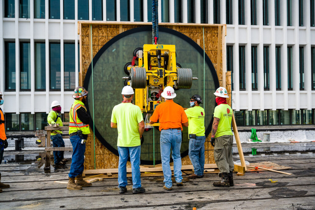 Construction crew guides a vacuum lift machine positioning a large circular glass floor panel at a Houston commercial jobsite.
