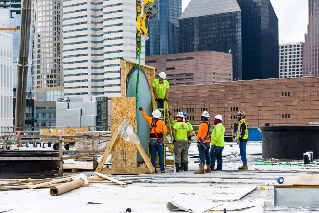 Construction crew installs a large oval glass flooring panel via crane lift on a rooftop jobsite in downtown Houston, Post HTX project.