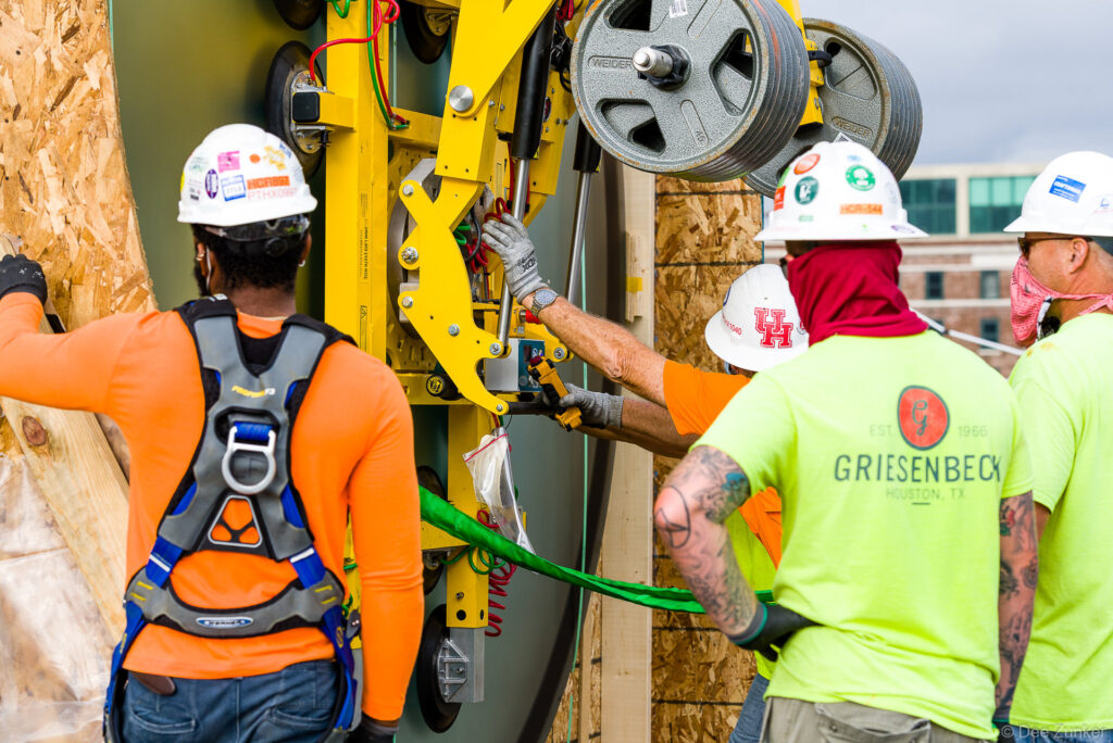 Construction workers operating a yellow vacuum lift system to install large glass panels on a Houston commercial jobsite, wearing hard hats and safety harnesses.