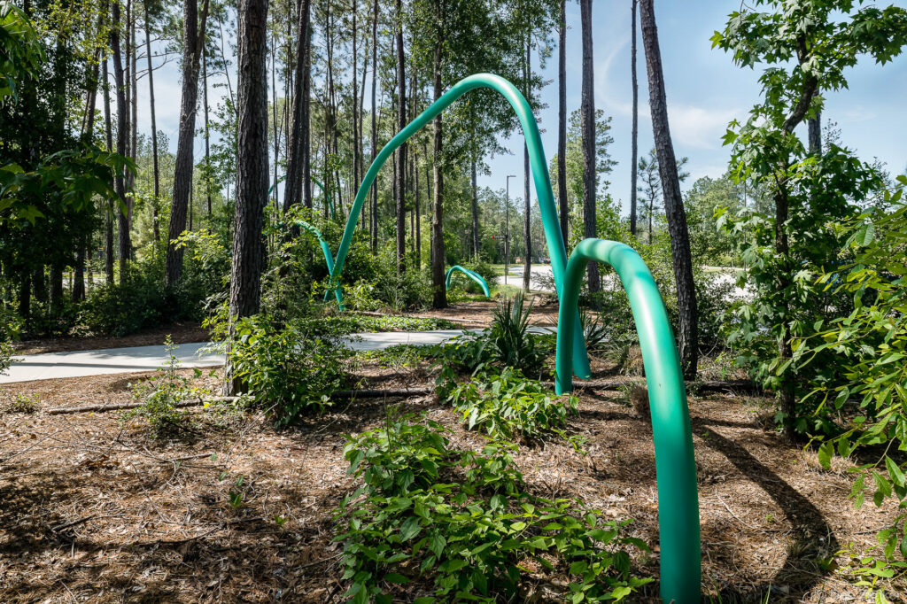 Teal green curved pipe sculptures arc through a wooded pine forest landscape with mulched garden beds and concrete path, Houston TX.