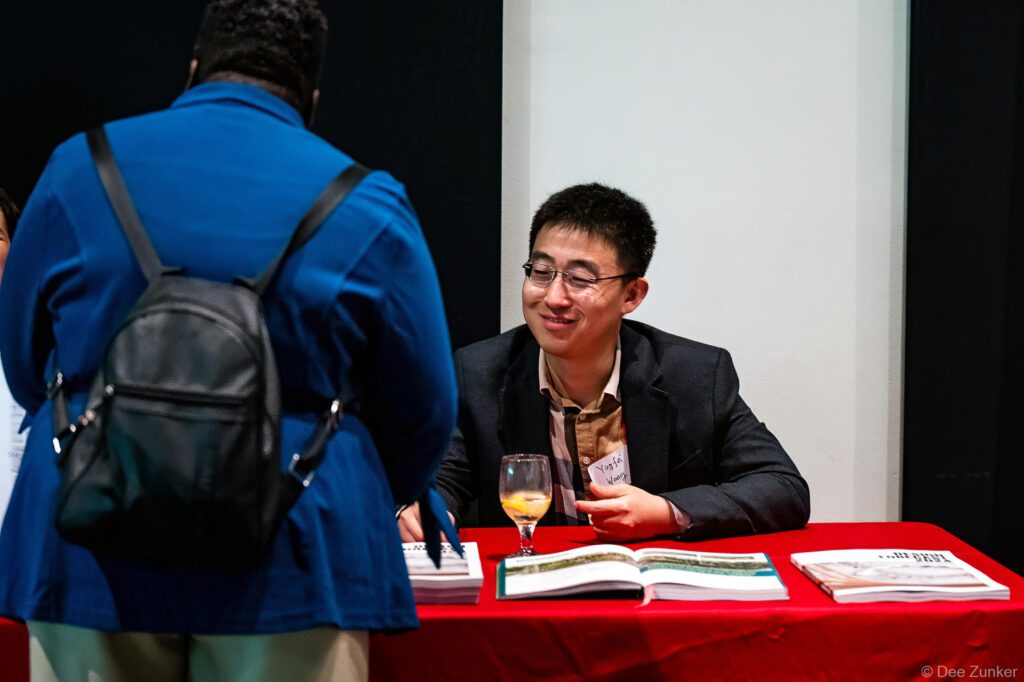 Young man in blazer holds wine glass at red-draped book signing table during Gulf Coast Green conference in Houston.