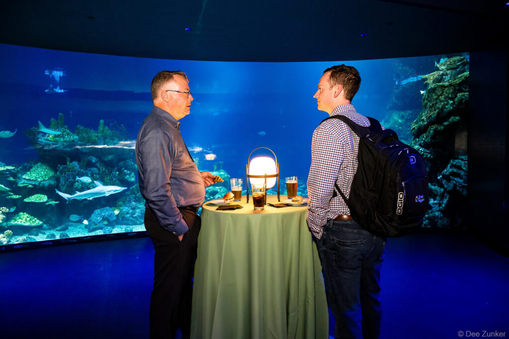 Two professionals networking at a cocktail table during Gulf Coast Green corporate event, with illuminated aquarium wall backdrop in Houston.