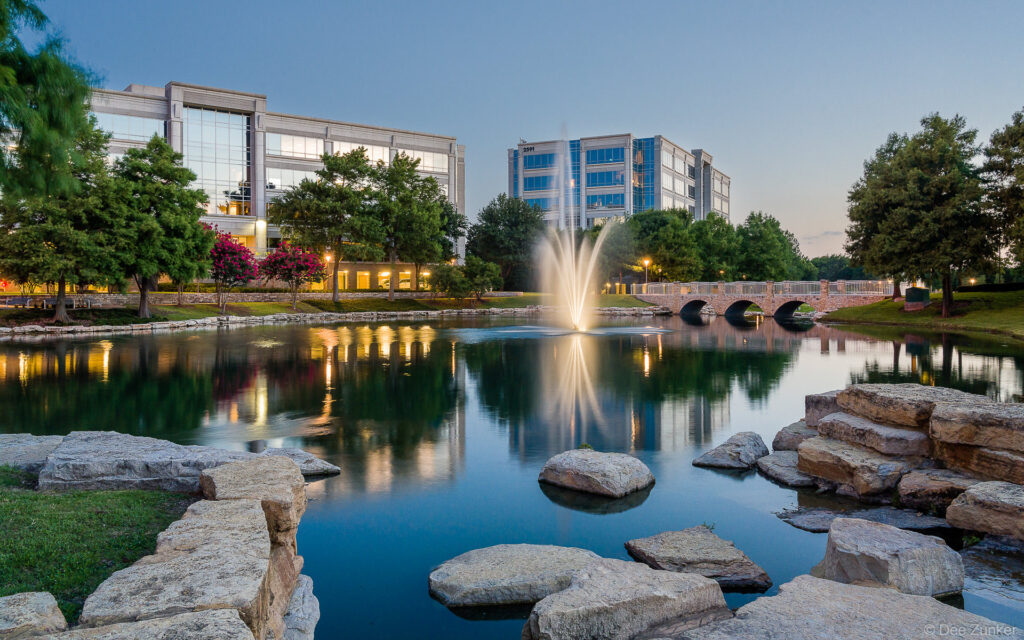 Illuminated fountain reflects in calm pond at Hall Park central plaza during twilight, Frisco Texas, professional landscape photography
