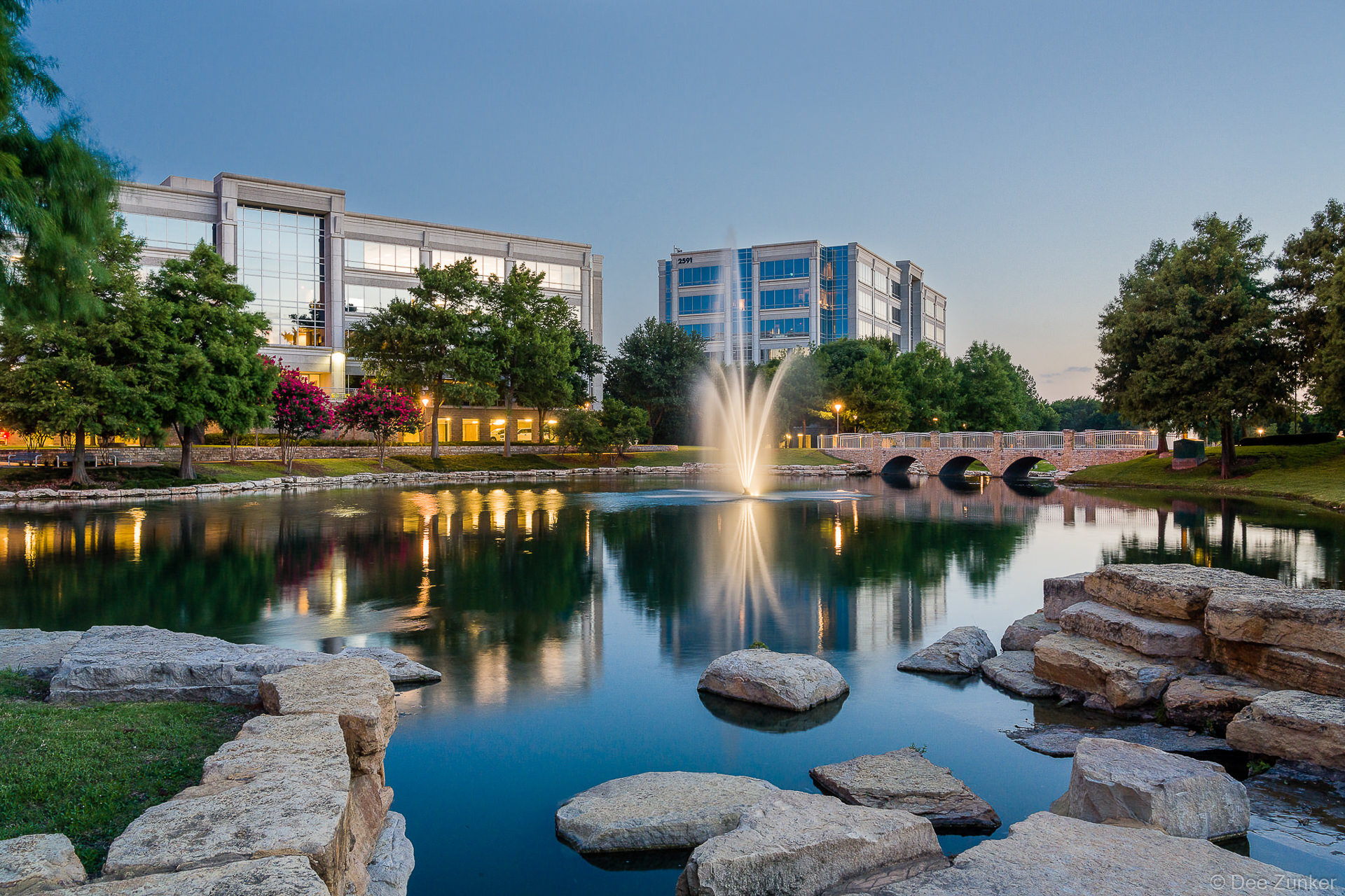 Illuminated fountain reflects in calm pond at Hall Park in Frisco TX at twilight with modern office buildings and stone bridge