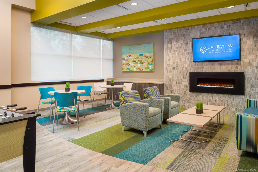Bright healthcare facility day room with teal and green seating, electric fireplace, flat screen TV, and foosball table in The Woodlands TX