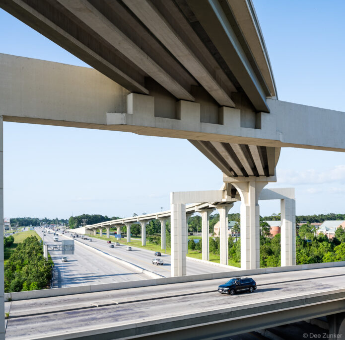 Underside view of elevated concrete highway interchange on Highway 99 in Houston, TX, with multilane traffic below and green surroundings.