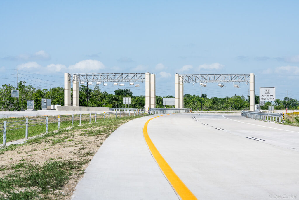 All-electronic toll gantry on Highway 99 Grand Parkway near Houston, TX, showing overhead sensor arrays and pay-by-mail signage on a concrete roadway.