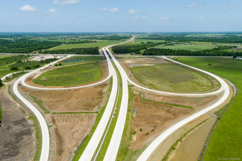 Aerial drone view of Highway 99 interchange construction showing sweeping concrete ramps and connector roads across open Texas landscape.