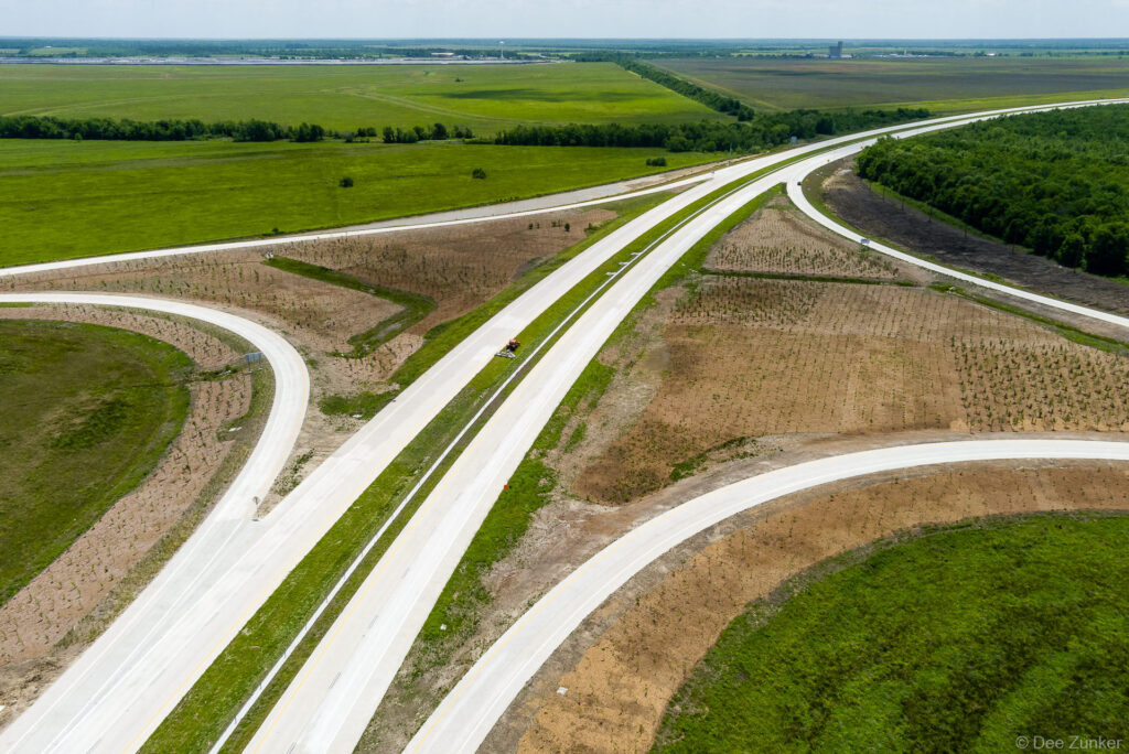 Aerial drone view of a newly constructed Highway 99 interchange with sweeping on-ramps, concrete lanes, and flat Texas farmland stretching to the horizon.