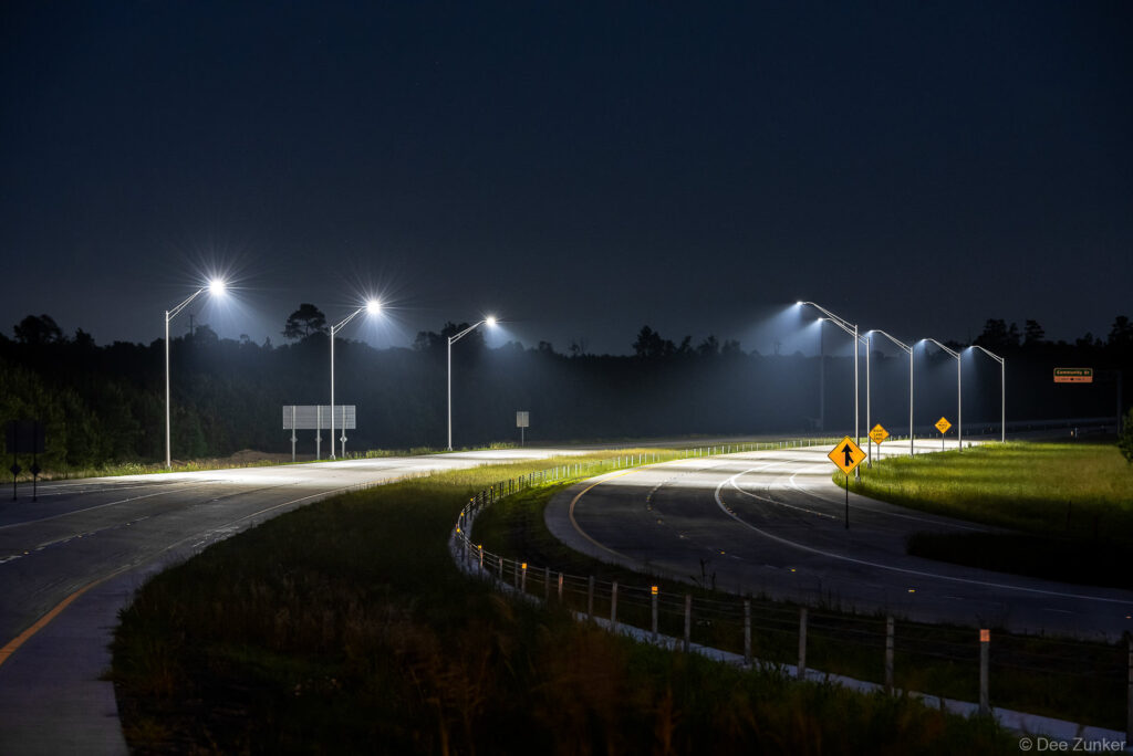 Illuminated Highway 99 interchange at night with glowing LED streetlights, curved on-ramps, and atmospheric mist over Texas infrastructure