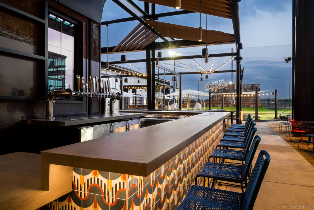 Outdoor bar interior at twilight featuring decorative tile facade, draft beer taps, blue bar stools, string lights, and open views to a green field in Katy Texas