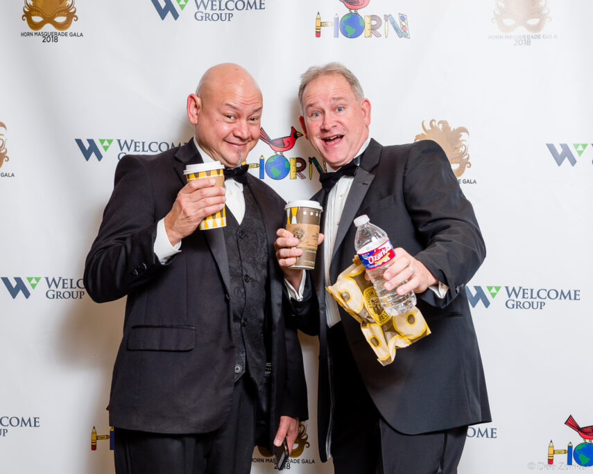 Two tuxedo-clad men pose playfully holding coffee cups and snacks at the Horn Masquerade Gala Houston corporate event.