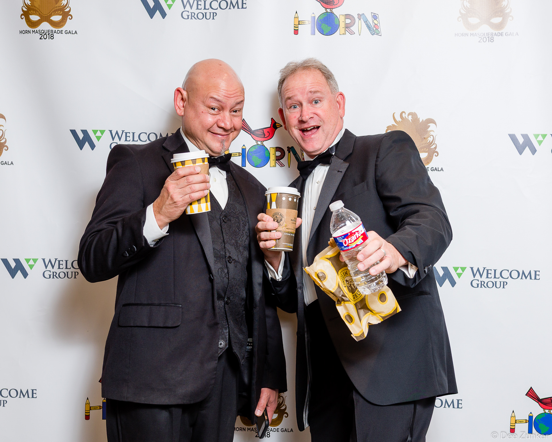 Two tuxedo-clad men pose playfully holding coffee cups and snacks at the Horn Masquerade Gala Houston corporate event.