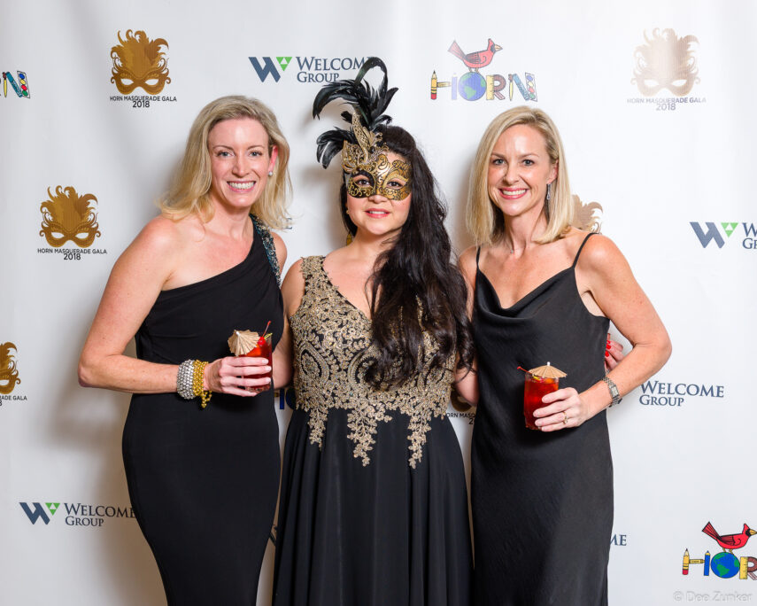 Three women in black formal gowns pose at Houston masquerade gala step-and-repeat backdrop, one wearing an ornate feathered mask.