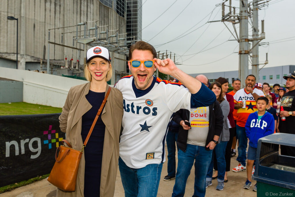 Two smiling Houston Astros fans in jerseys and cap pose outside NRG Park during the Domecoming corporate event.