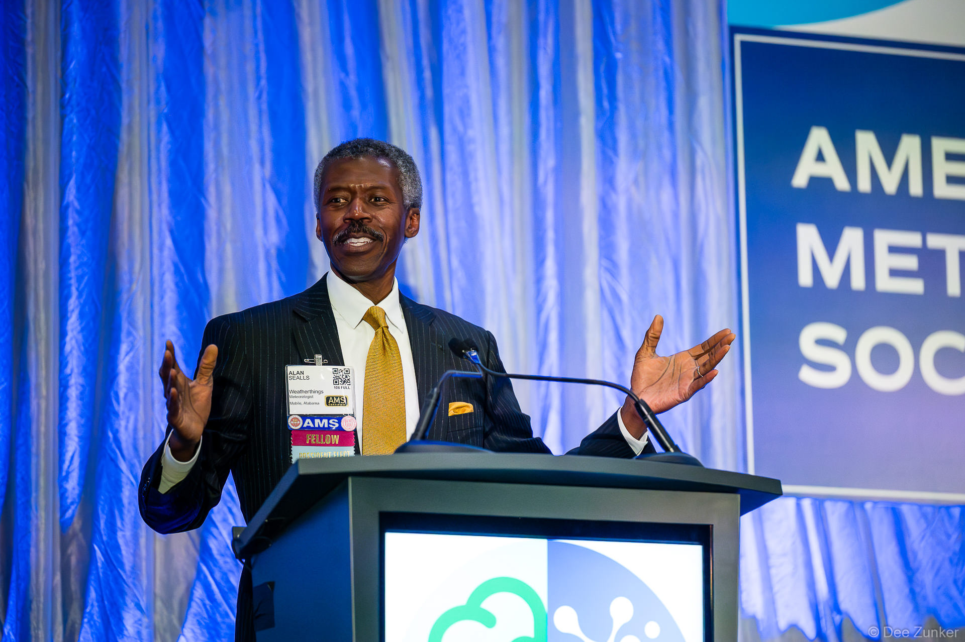 Keynote speaker at Houston awards ceremony gestures expressively at podium before professional meteorological society audience