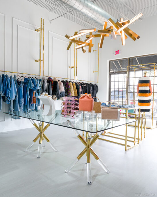 Upscale Houston boutique interior with glass display table, gold lighting fixture, handbags, sunglasses, and clothing racks with denim and fur coats.