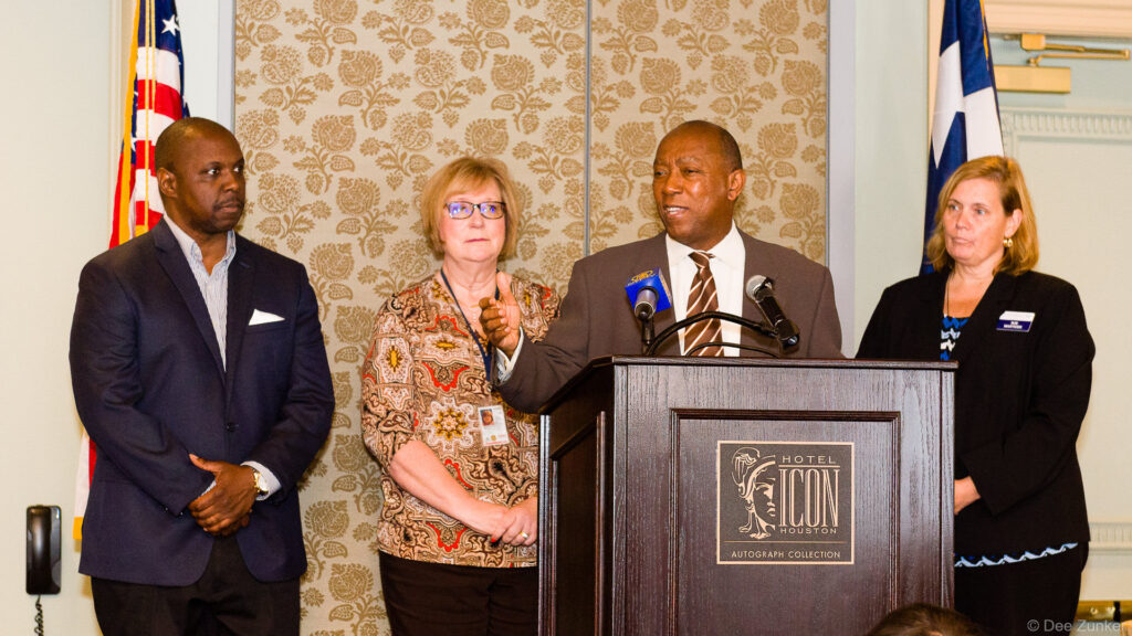 Speaker at Hotel Icon Houston podium addresses three colleagues during a Houston chamber corporate event, Houston TX.