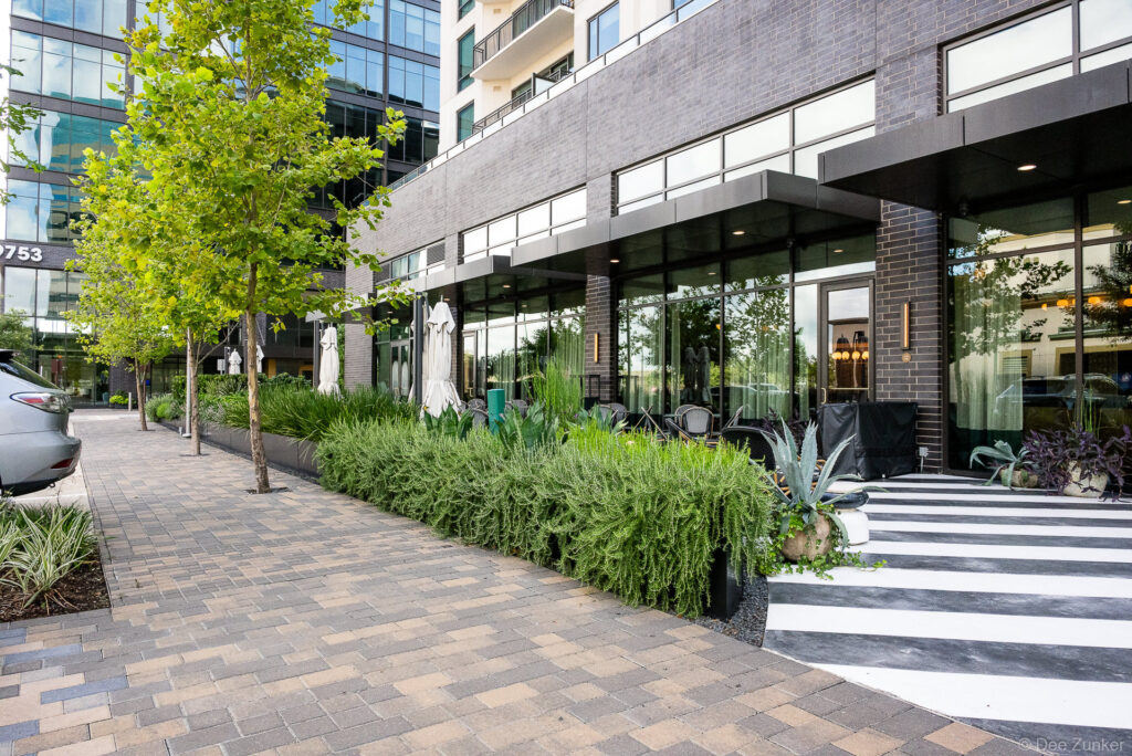 Lush landscaped courtyard beside a dark brick commercial building in Houston, featuring ornamental grasses, agave, and paver walkway.