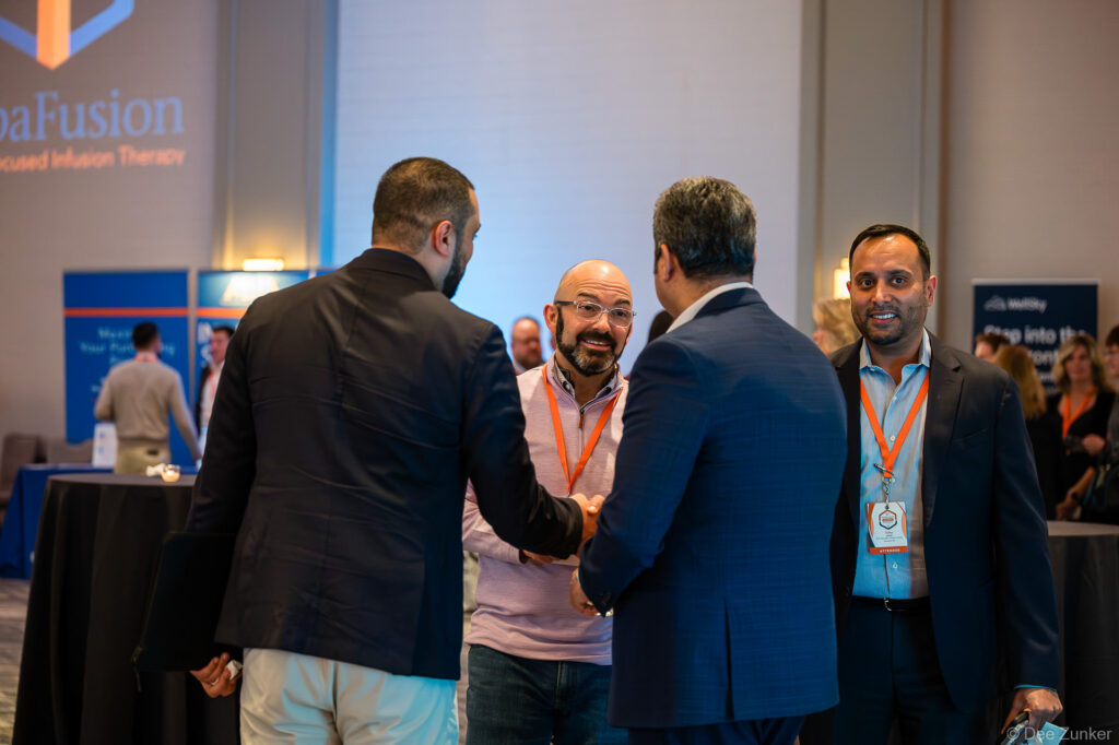 Conference attendees networking and shaking hands at a Houston corporate event, with branded signage visible in the background