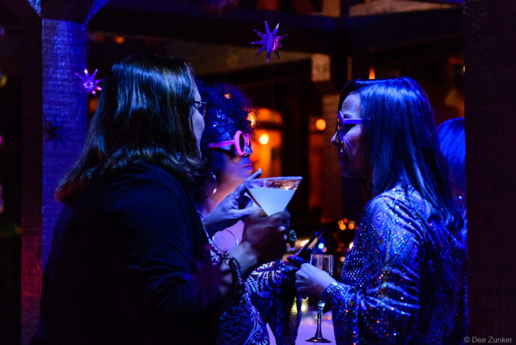 Three women socializing at a Houston corporate 40th anniversary evening event, holding cocktails under purple star decorations in dim blue lighting.