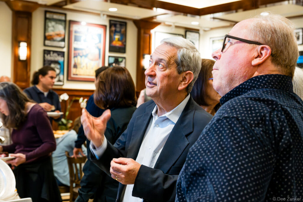 Two older men conversing at an indoor Houston corporate birthday celebration event, with guests mingling and framed artwork on wood-paneled walls.