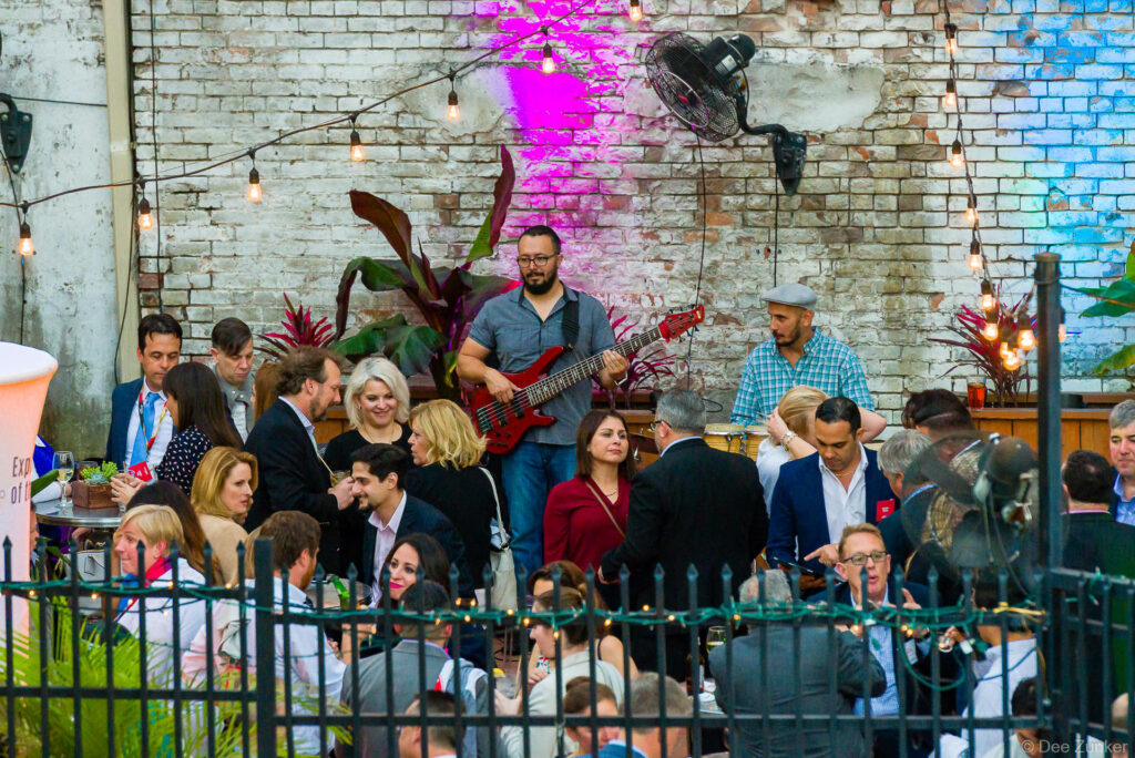Guitarist performs live at a crowded Houston corporate event reception in a brick courtyard with string lights and tropical plants.