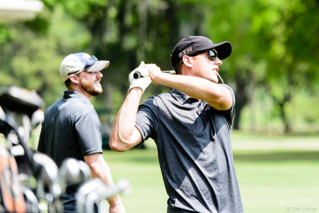 Two male golfers at a Houston corporate golf event, one mid-swing follow-through wearing an RZN cap and sunglasses, another watching nearby.