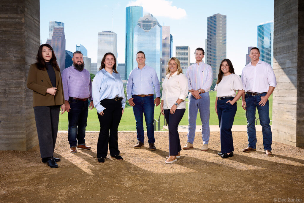 Eight corporate professionals posed outdoors at Buffalo Bayou Park with the Houston downtown skyline in the background, corporate group portrait.