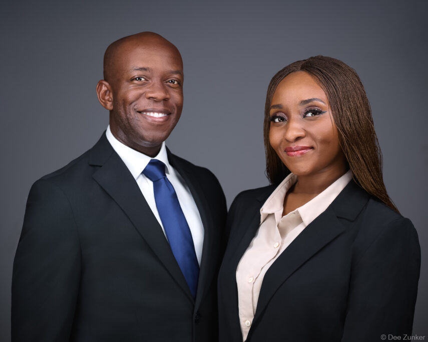 Two professionals in dark business suits pose for a corporate group headshot in a Houston photography studio with gray background.