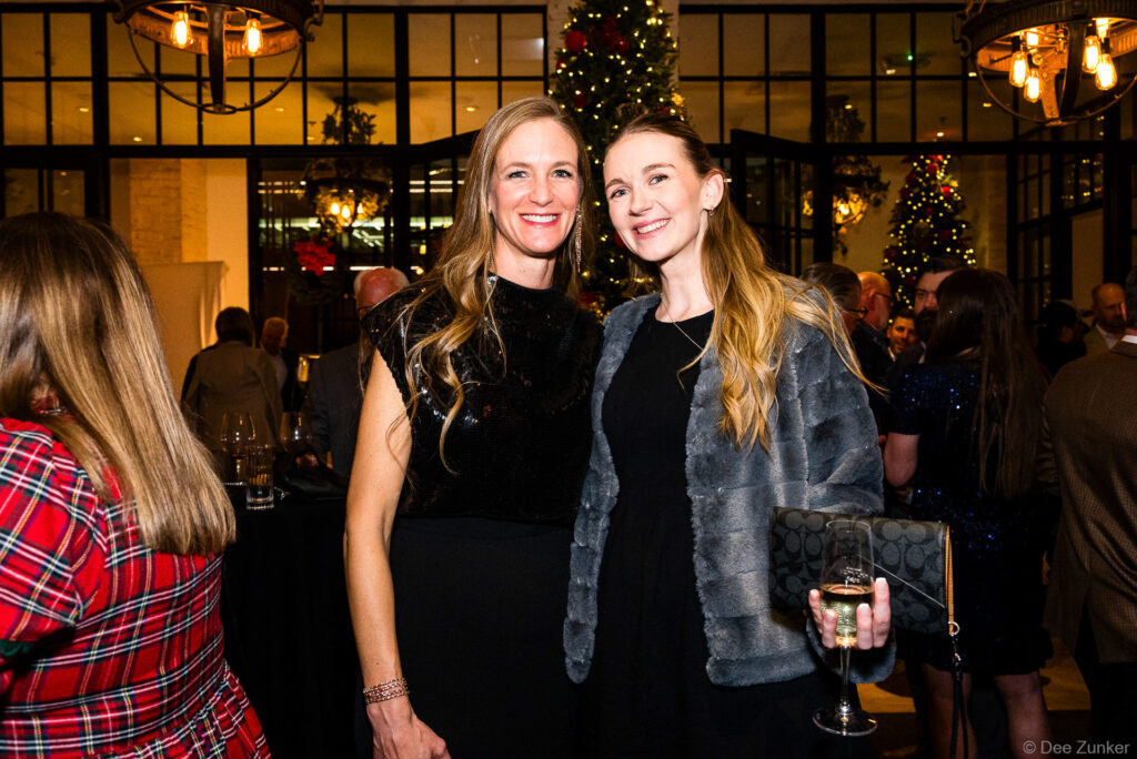 Two women smiling together at a Houston corporate Christmas party, decorated tree and warm event lighting in background