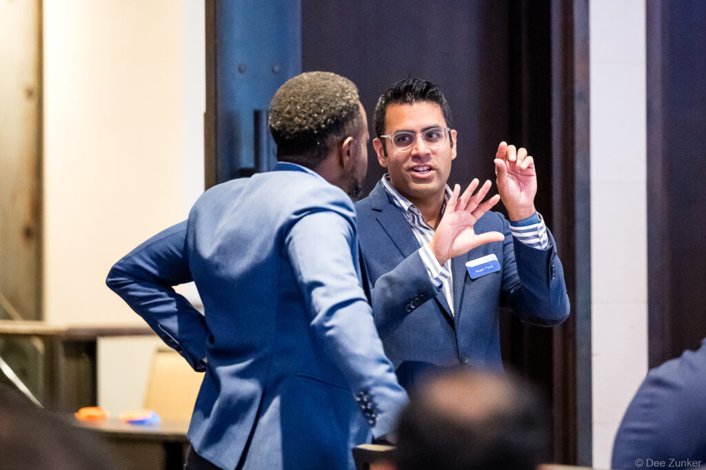 Two professionals in navy blazers engage in animated conversation at a Houston corporate networking event, one gesturing expressively.