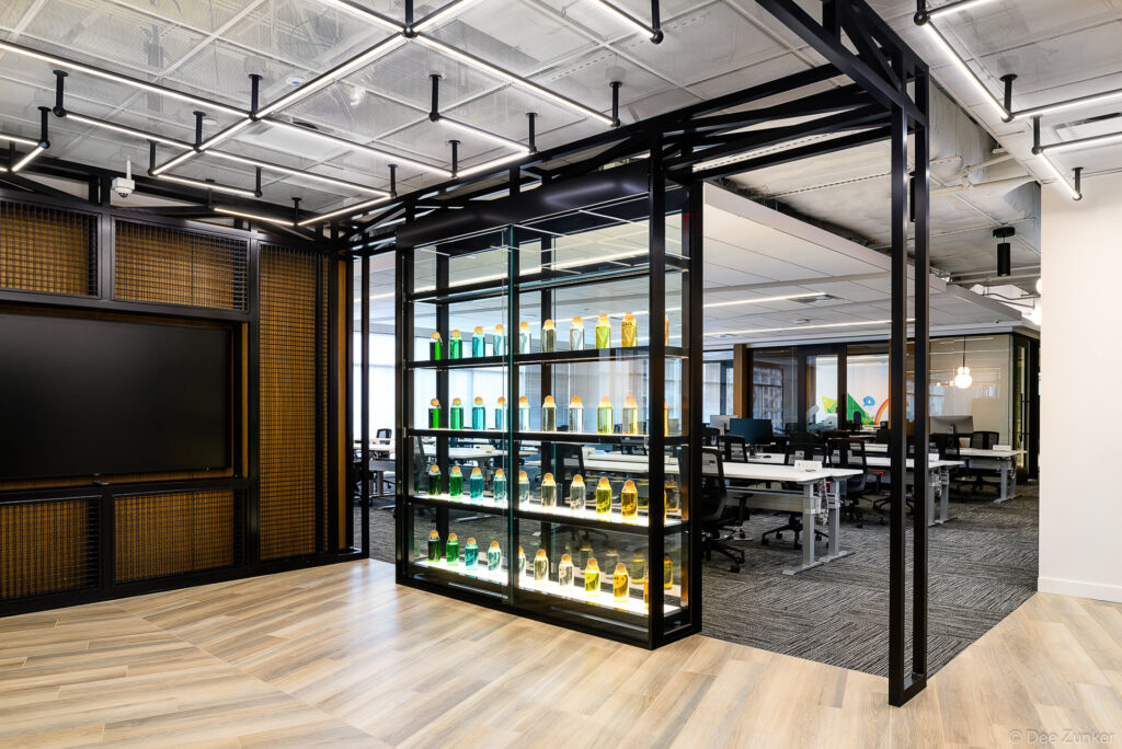 Modern corporate office interior with glass display shelving, colorful product bottles, open workstations, and industrial black metal framework