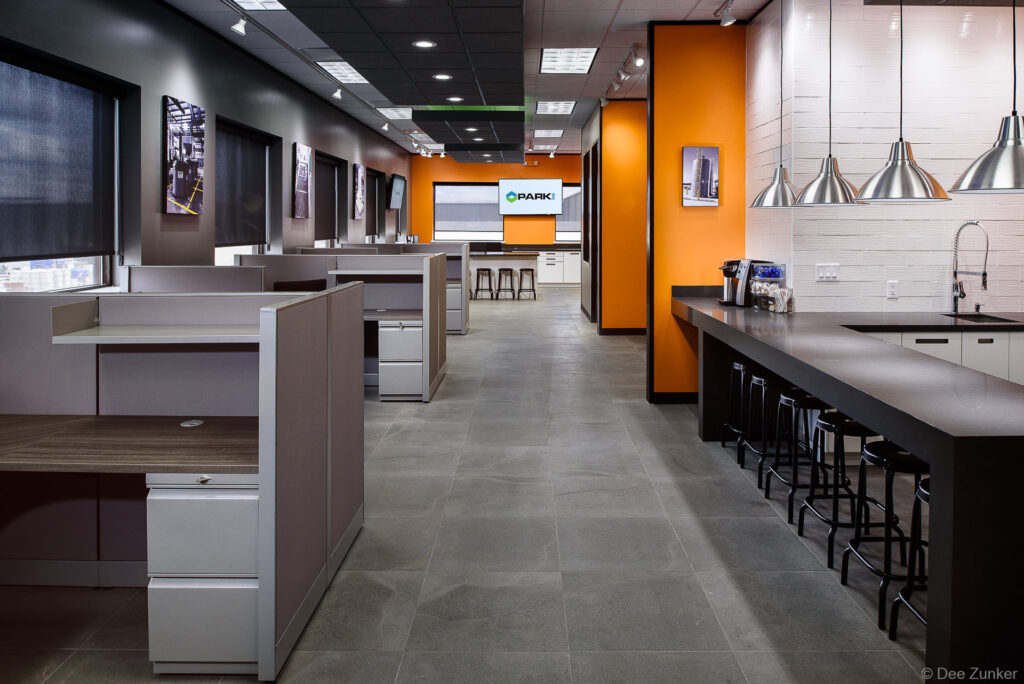 Open-plan corporate office interior with cubicle workstations, dark kitchen bar seating, orange accent wall, and branded display screen in Houston.