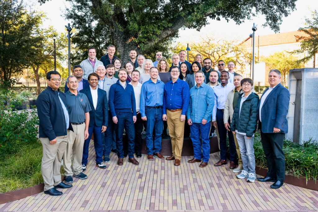 Large corporate team group photo outdoors in Houston, approximately 30 professionals posed on a brick plaza beneath a large oak tree.
