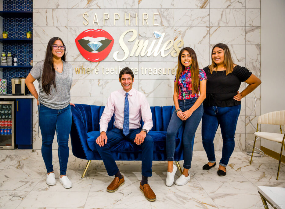 Dental office team of four staff members pose in modern marble-tiled reception area with blue velvet sofa, Houston, TX.