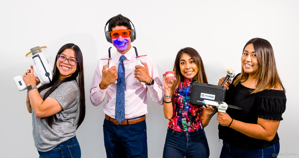 Four smiling dental professionals pose with dental equipment including an Invisalign case and curing light in a Houston studio.