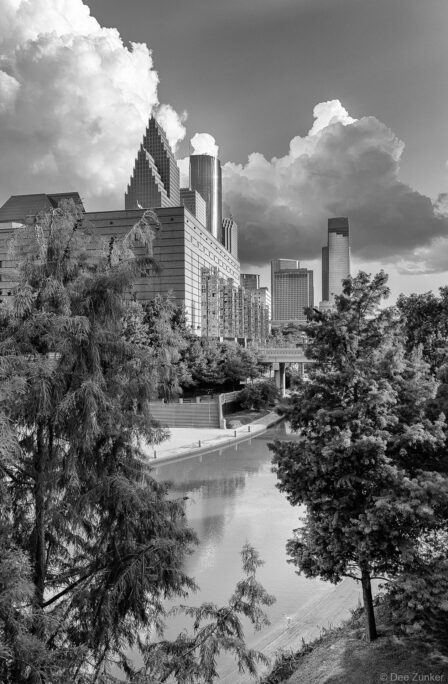 Black and white aerial view of Houston downtown skyline with Buffalo Bayou canal, trees, and dramatic cloudy sky, architectural exterior photography.