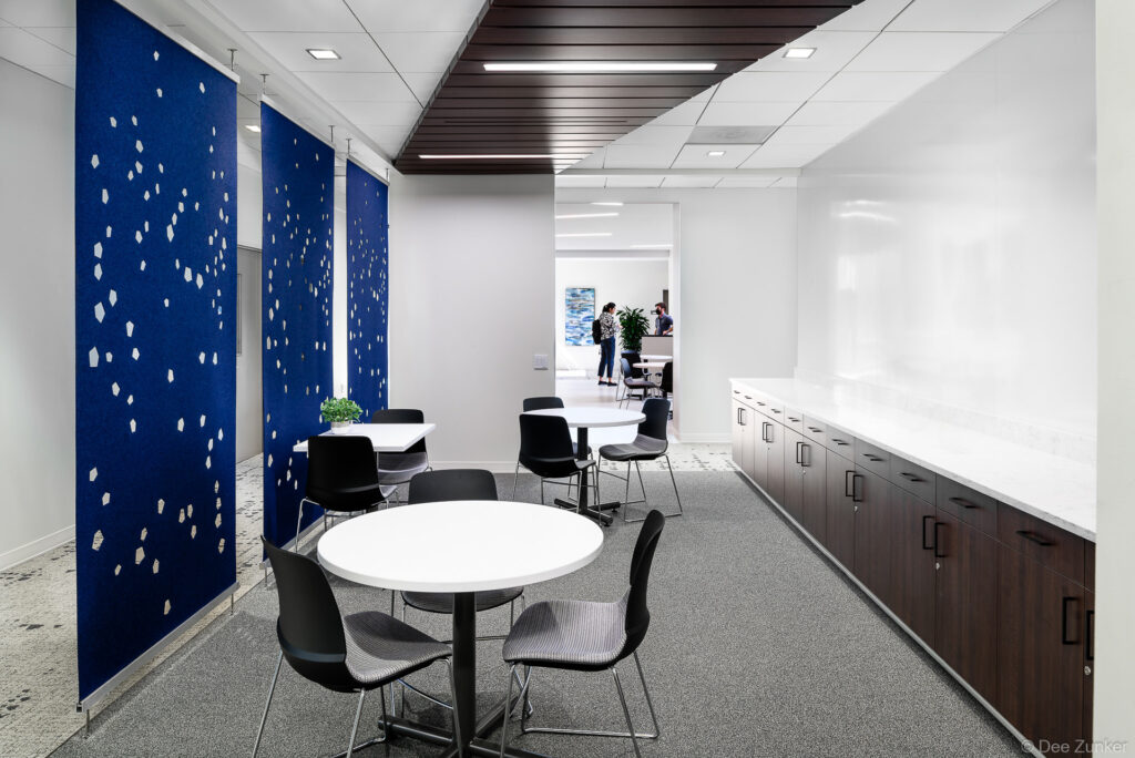 Renovated Houston office breakout area with round white tables, black chairs, blue perforated acoustic panels, and dark wood cabinetry