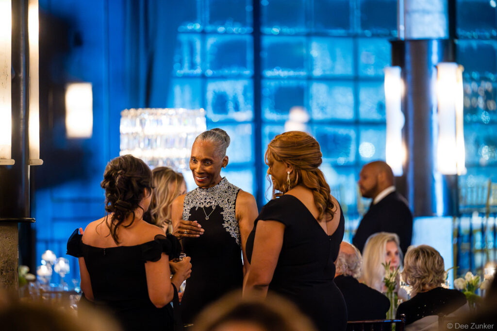 Elegantly dressed guests networking at a Houston corporate gala event, candid moment captured in a glamorous blue-lit venue