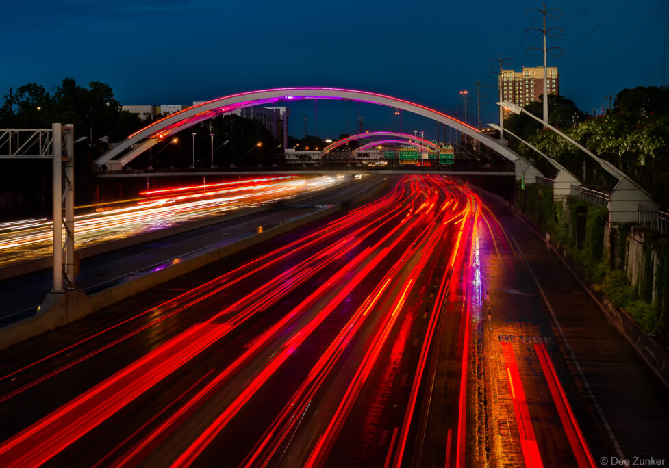 Long-exposure night photography of Houston Highway 59 showing red light trails beneath illuminated arch bridges, reflecting on wet pavement