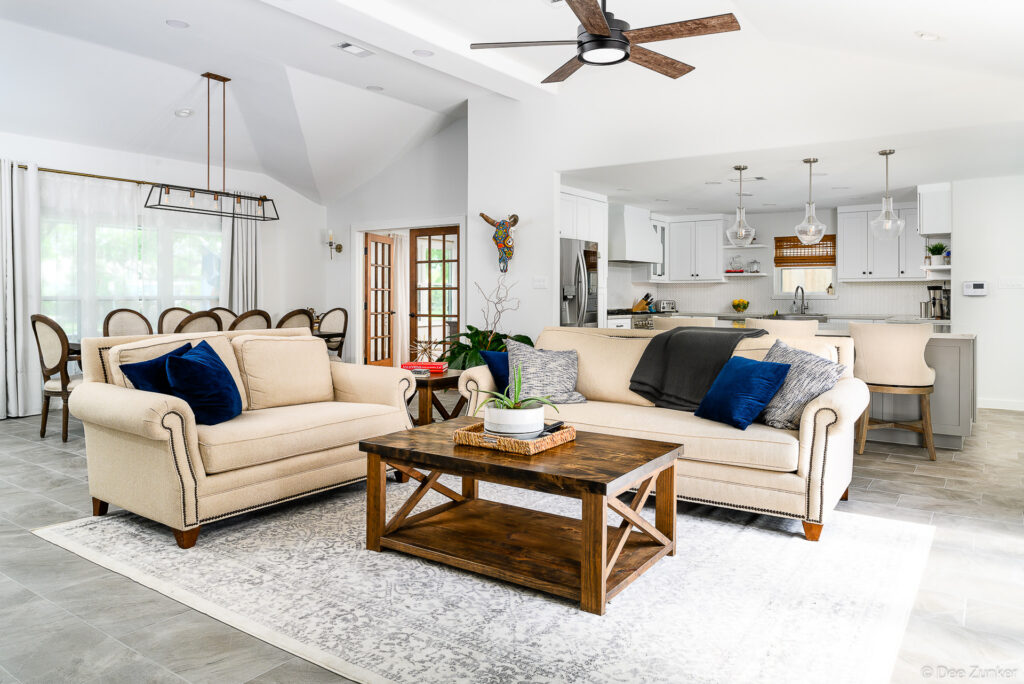 Bright open-concept living room with cream sofas, blue velvet pillows, rustic wood coffee table, and white kitchen visible beyond, Houston residential interior.
