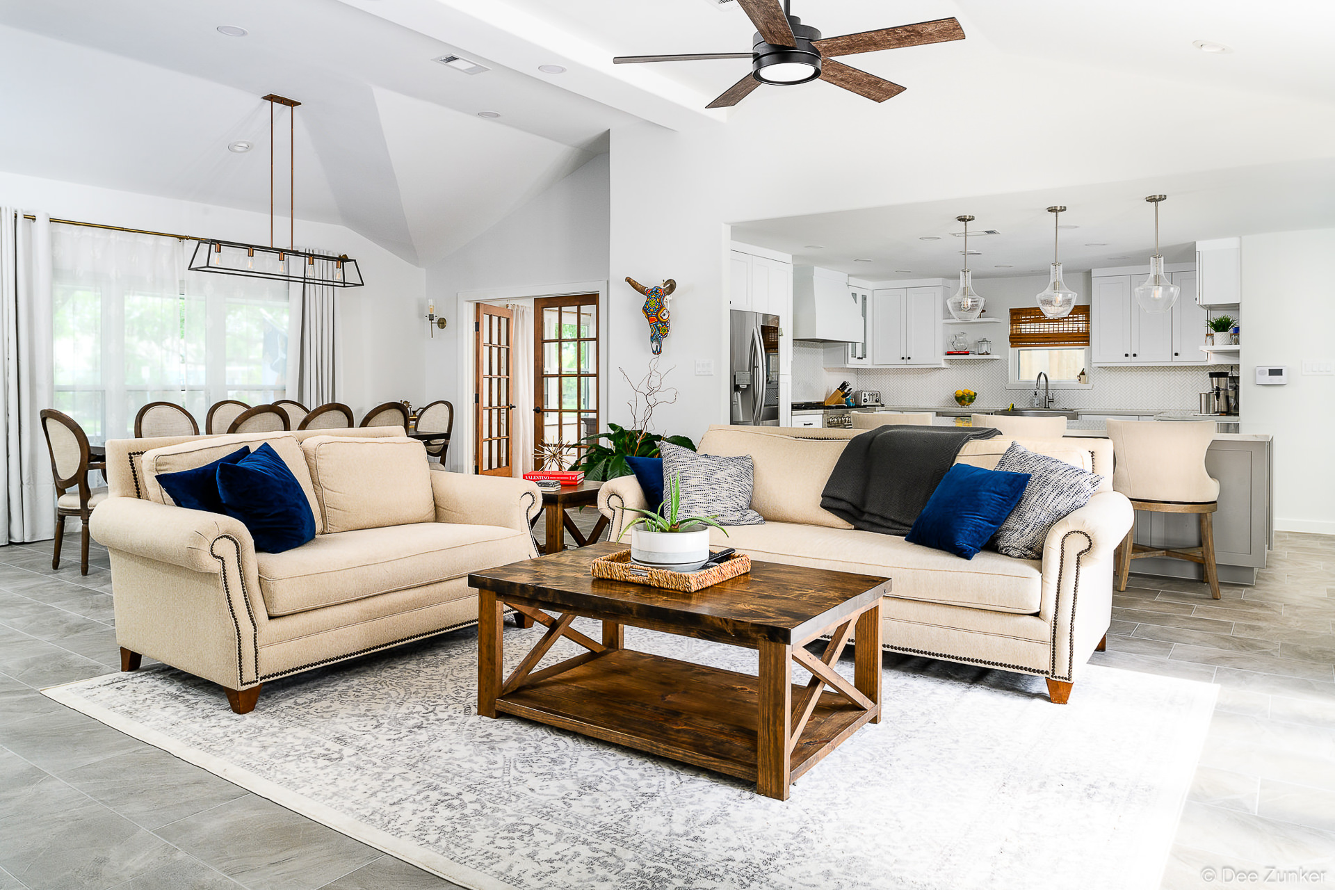 Bright open-concept living room with cream sofas, blue velvet pillows, rustic wood coffee table, and white kitchen visible beyond, Houston residential interior.